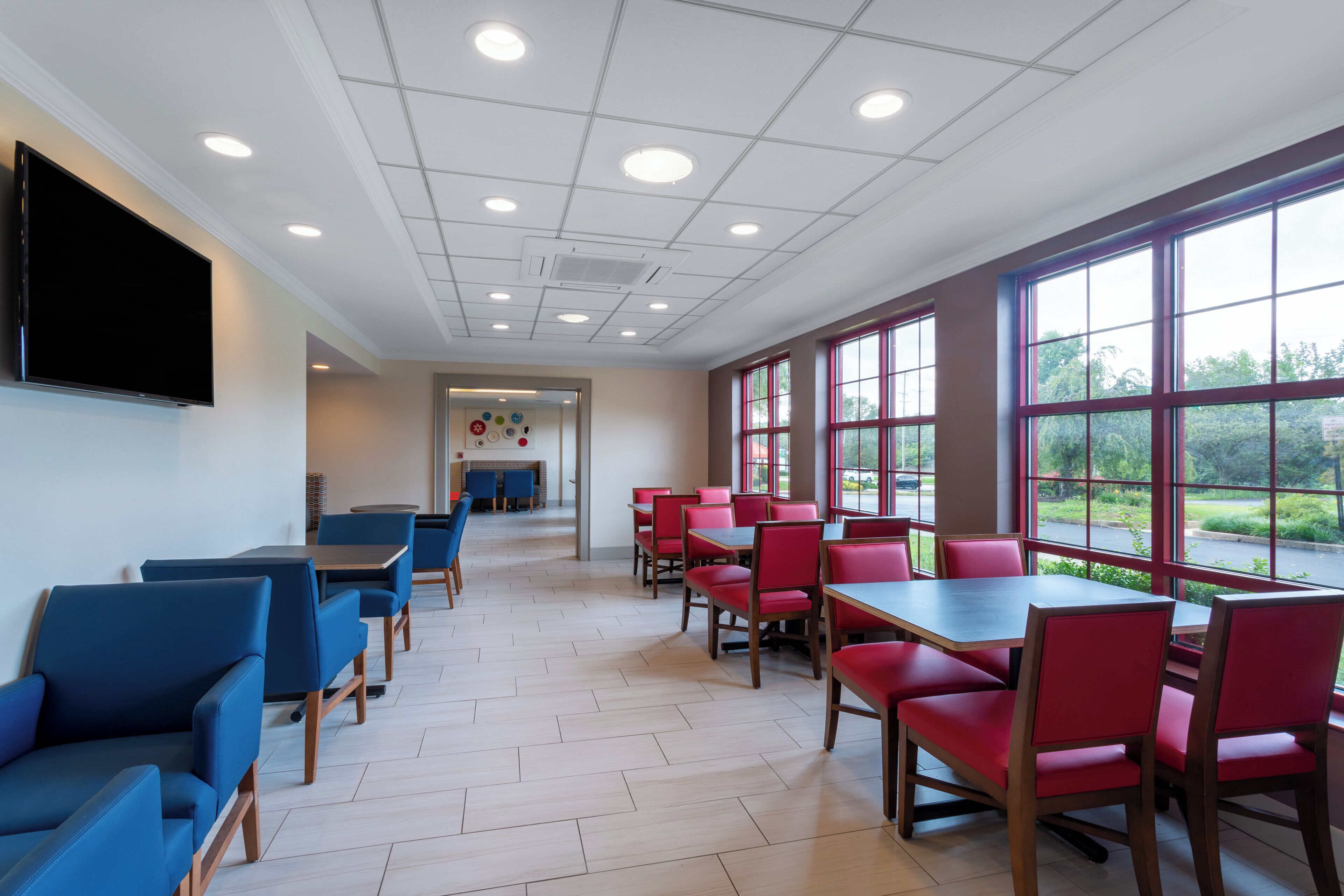 A dining area with tables and chairs, a flat-screen TV mounted on the wall, and large windows with a view of the outdoors.