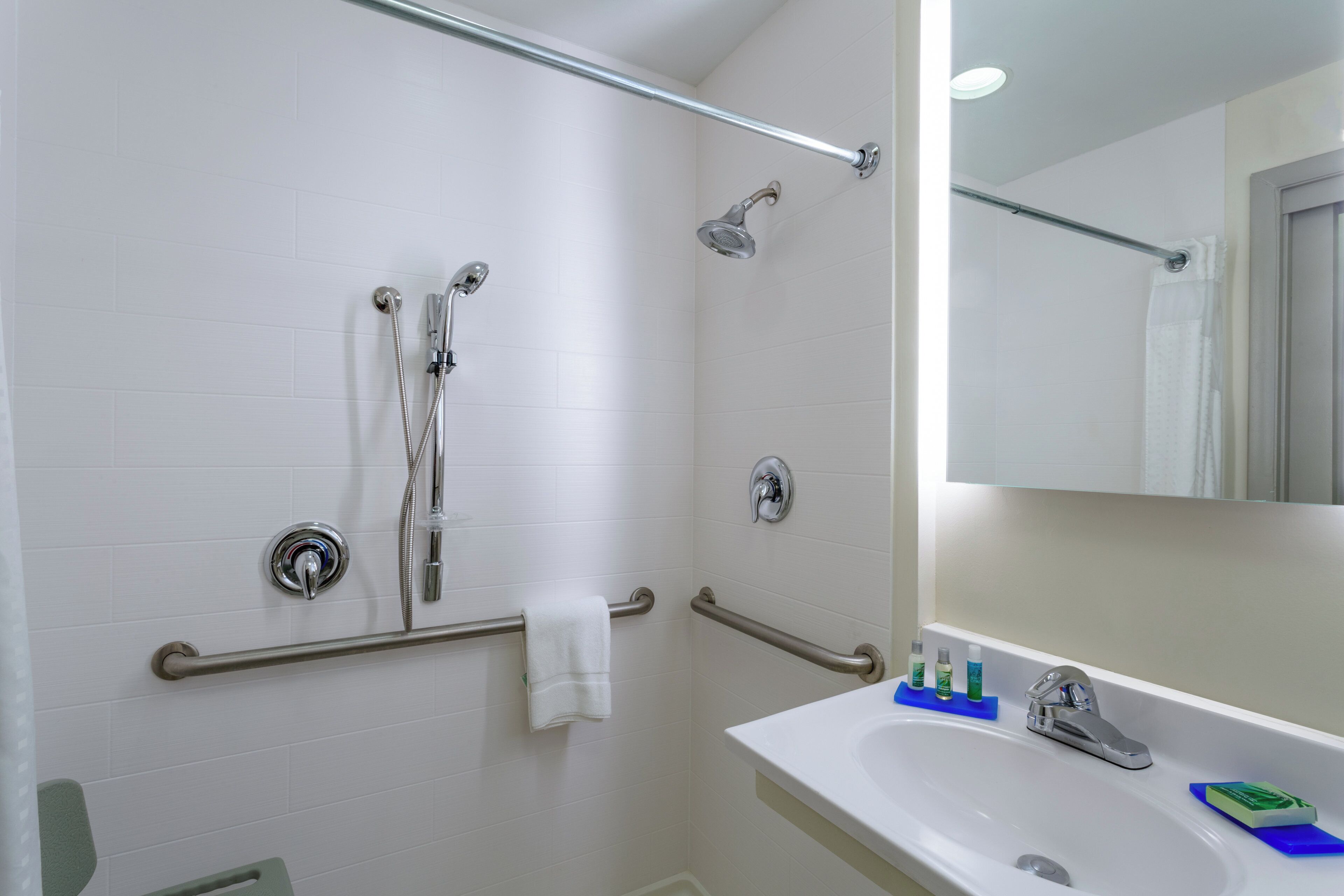 A bathroom with a shower, grab bars, a towel rack, a sink with toiletries, and a mirror.
