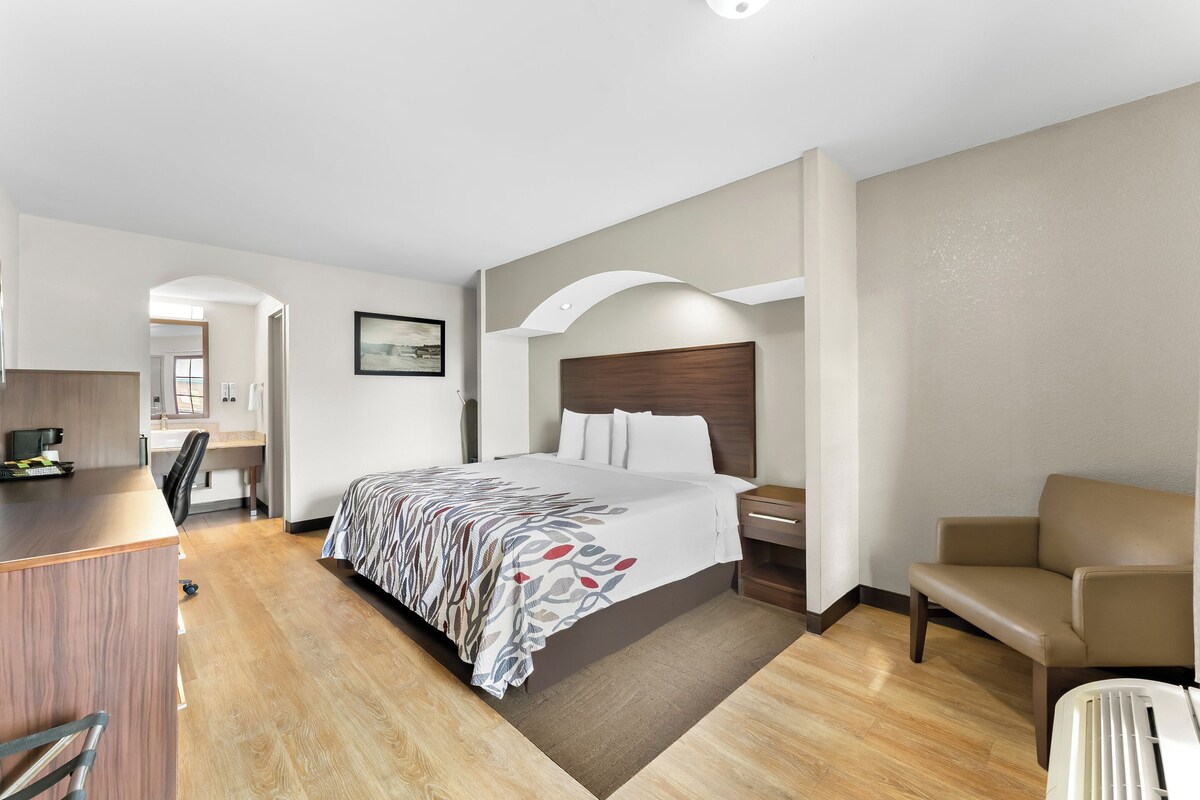 A hotel room with a bed, desk, chair, and a view of the hallway.