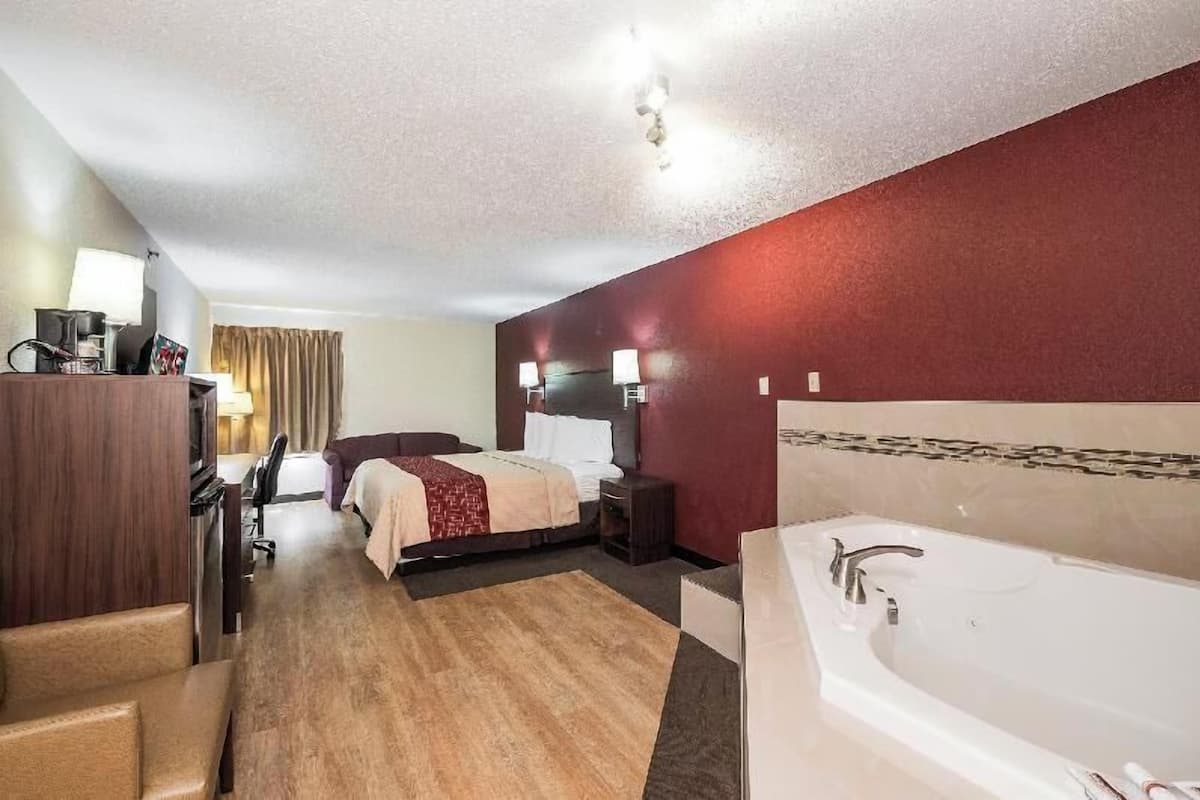A hotel room with a bed, a desk with a computer, a chair, a desk lamp, a TV, a minibar, a bathroom with a bathtub, and a red accent wall.
