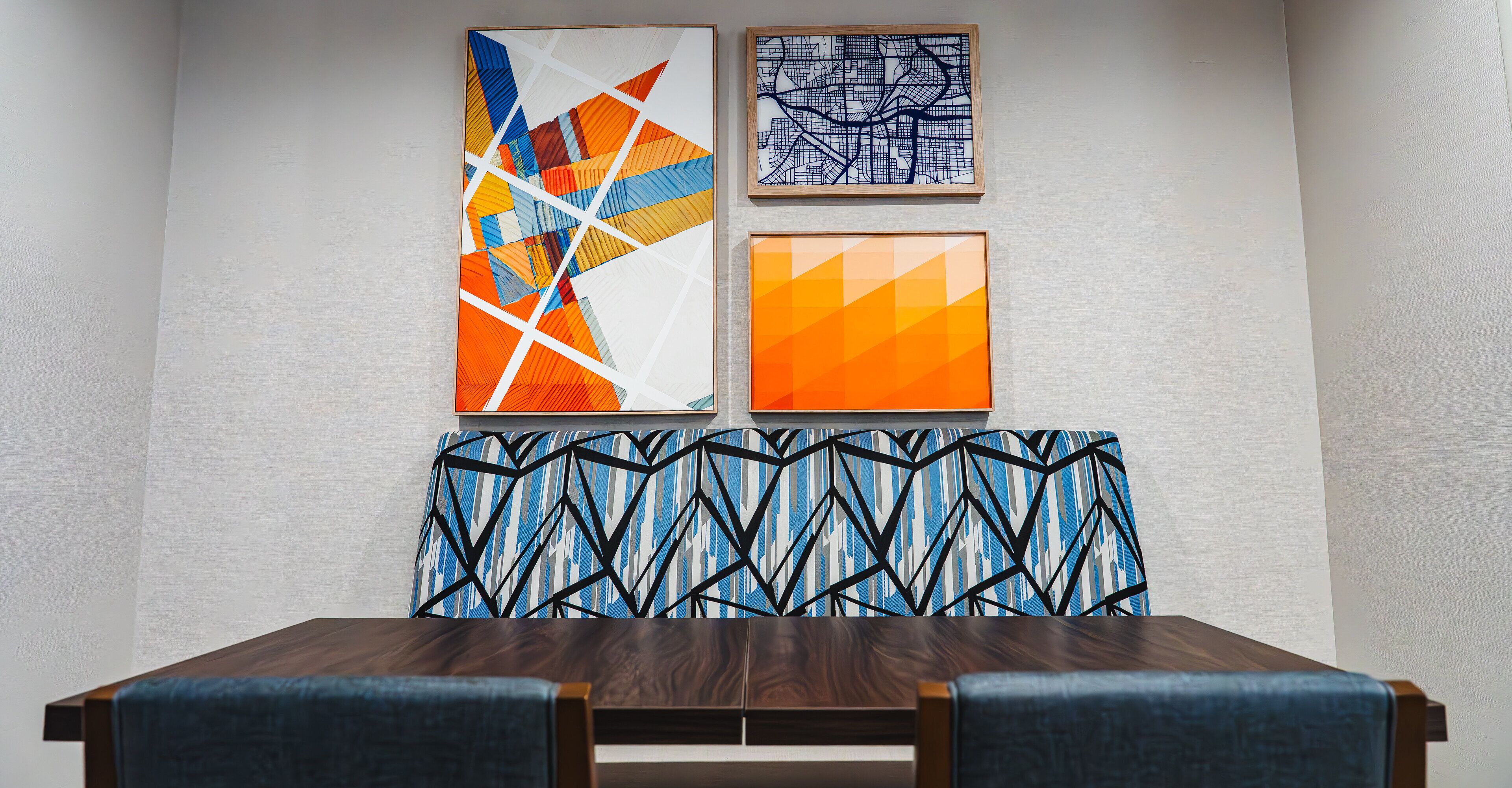 Zona pranzo con tavolo in legno e sedie rivestite in blu, pannello decorativo a parete e tre opere d'arte incorniciate.