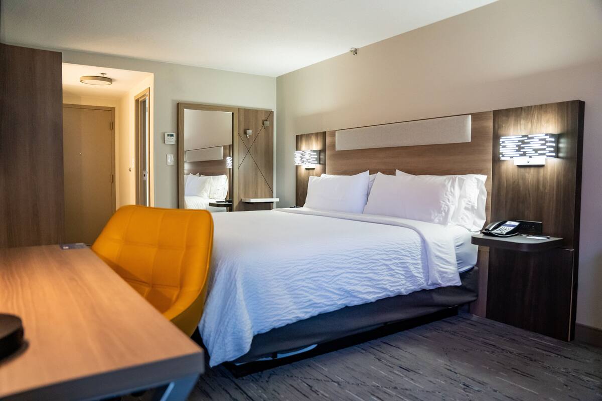 A hotel room with a bed, a desk, a chair, a nightstand, and a mirror.