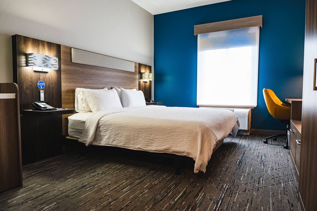 A hotel room with a bed, bedside table, lamp, and a window with blinds.