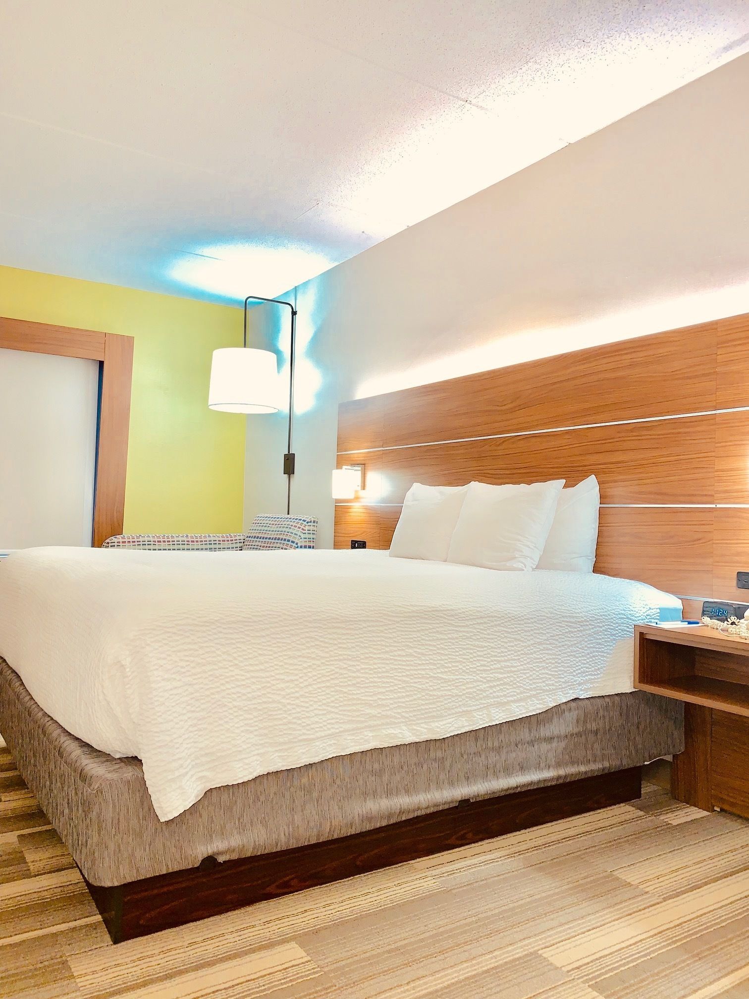 A hotel room with a large bed, a wooden headboard, a bedside table with a lamp, and a nightstand with a phone.