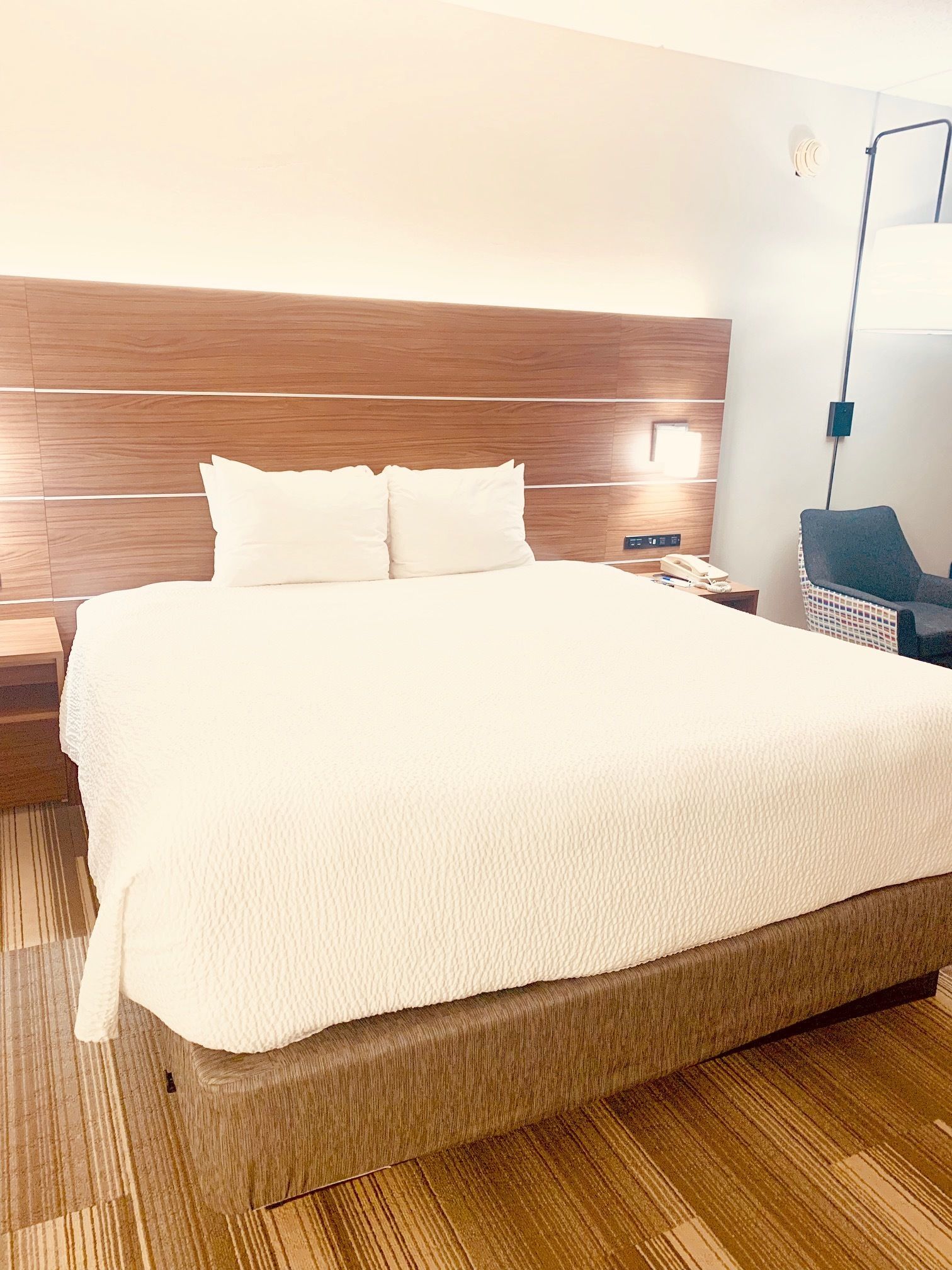 Una habitación de hotel con una cama grande, cabecera de madera, una mesita de noche con lámpara y una silla.