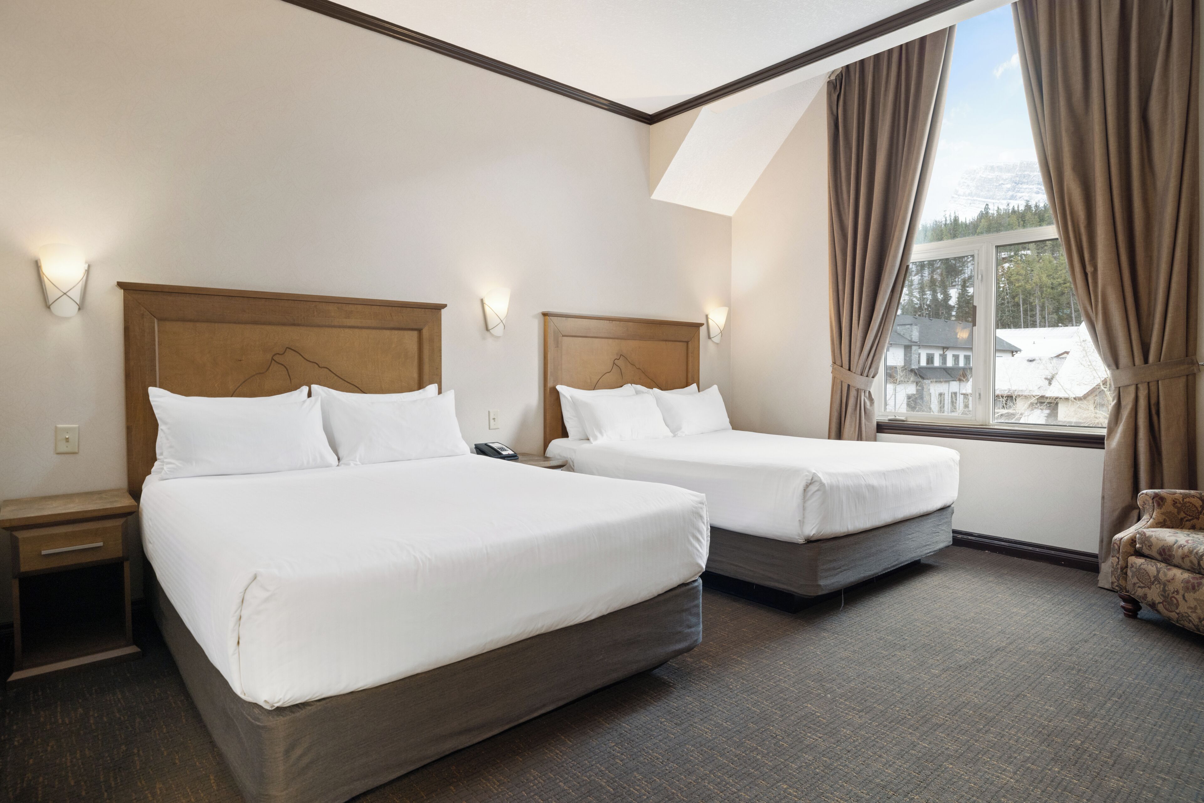 A hotel room with two beds, a small armchair, a window with curtains, and a view of a snowy landscape.