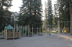 A playground with various play structures, surrounded by trees and a building in the background.