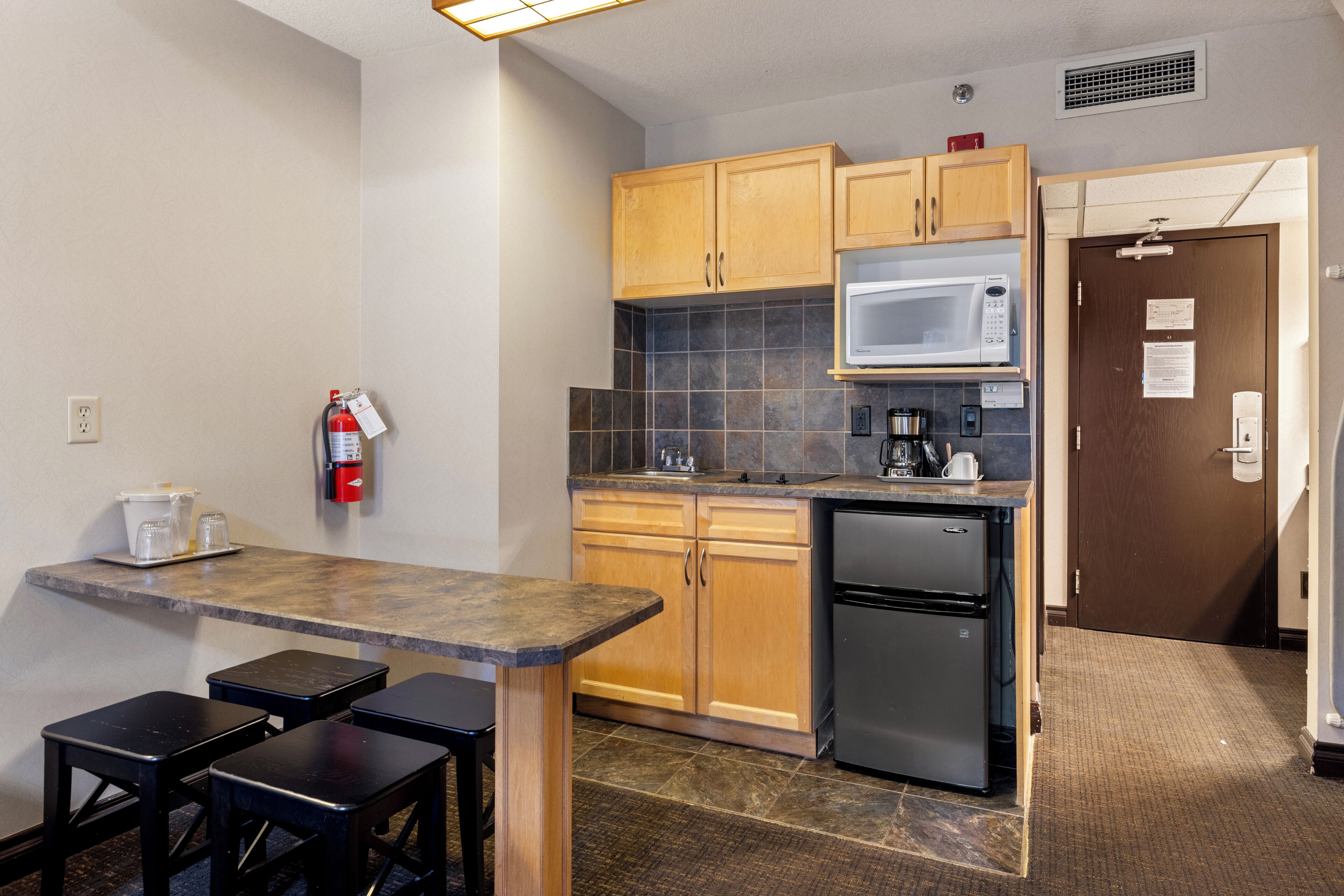A compact kitchen with wooden cabinets, a built-in microwave, a coffee maker, and a refrigerator. There is a small bar area with stools and a fire extinguisher mounted on the wall.