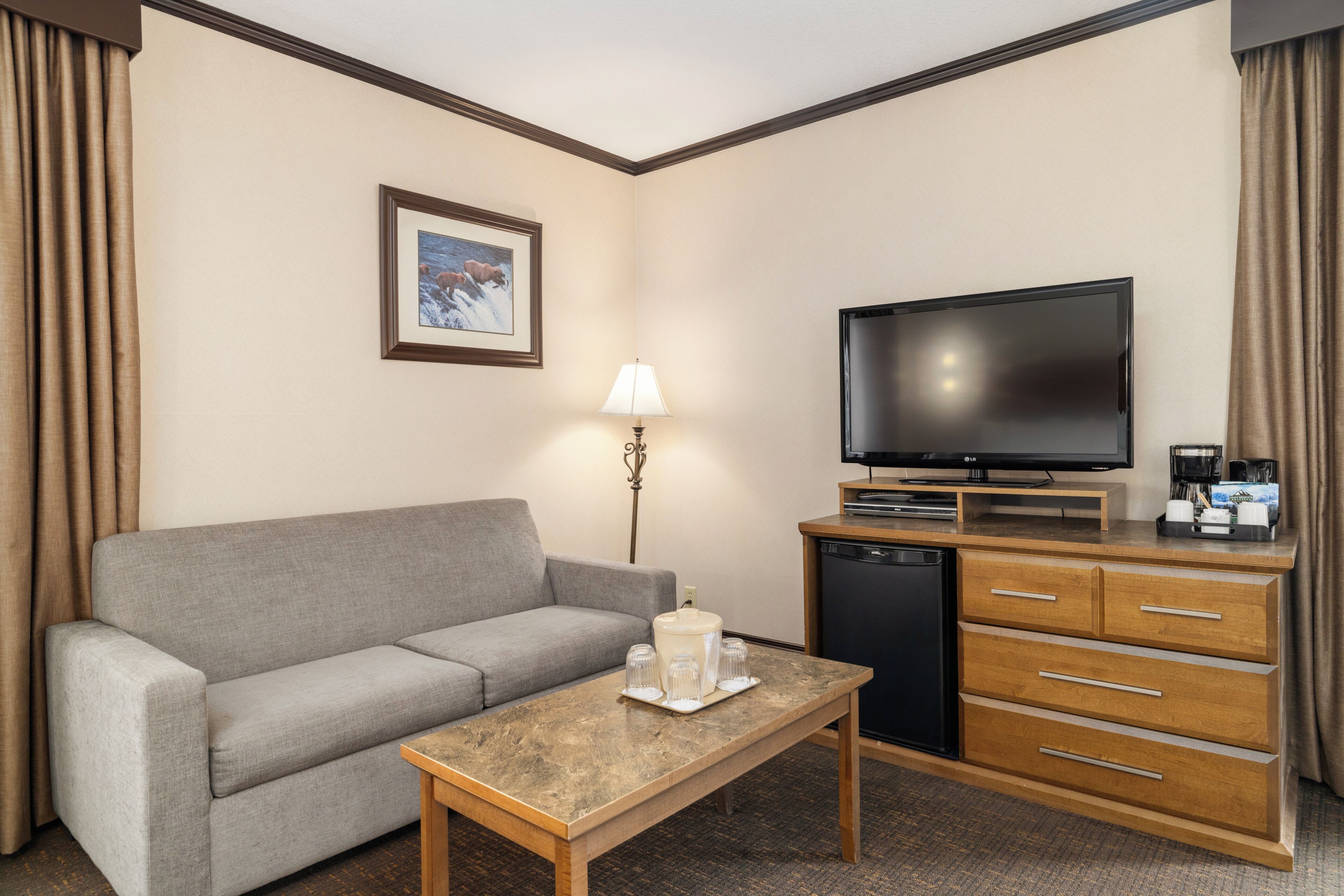 A hotel room with a grey sofa, a wooden coffee table, a flat-screen TV, a mini-fridge, a coffee maker, and a lamp.