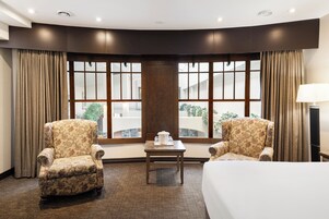 A hotel room with a large window, two armchairs, a small table, and a bed.