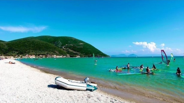 Una playa con agua turquesa cristalina, un bote en la orilla y gente practicando windsurf.