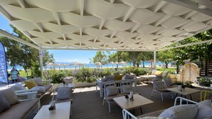 Un área de estar al aire libre con mesas y sillas, techo cubierto y vista a la playa y al mar.