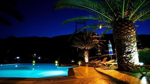 Zona de piscina nocturna con sillones de descanso y palmeras, iluminada con luces ambientales.