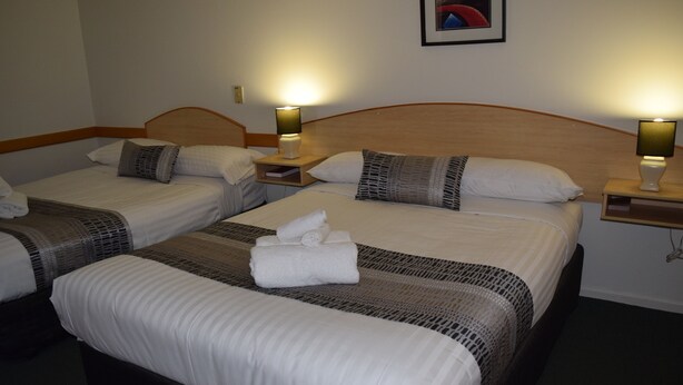 A hotel room with two beds, each with a wooden headboard, white linens, and a small bedside table with a lamp. - Warwick Vines Motel (Warwick)