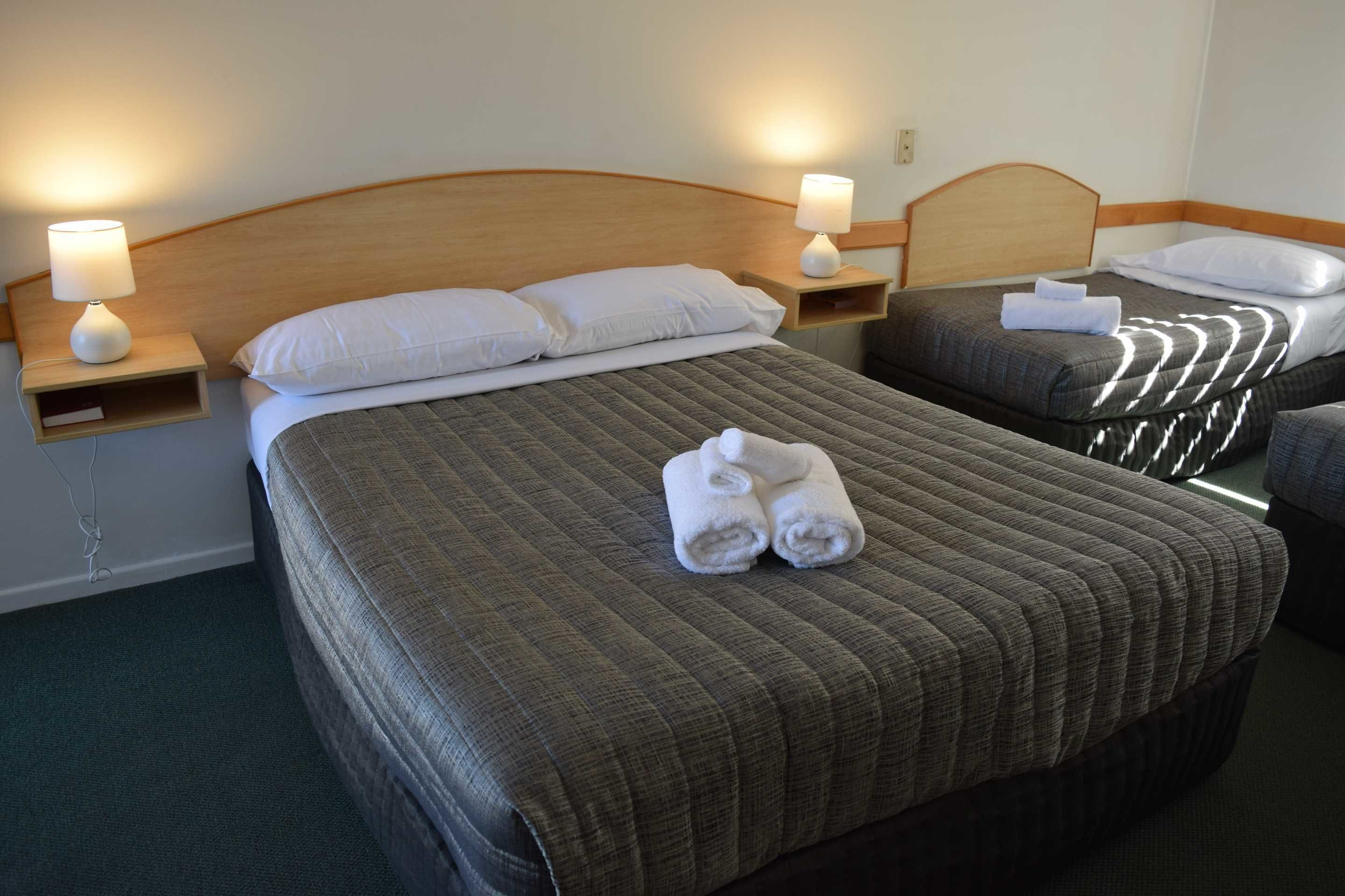 A hotel room with two beds, each with a wooden headboard, a bedside table with a lamp, and a pair of rolled towels on each bed.