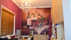 A compact room with a desk, a computer, a refrigerator, and a microwave. There is a window with blinds, a chandelier, and a wall mural depicting wine bottles and grapes. - Warwick Vines Motel (Warwick)