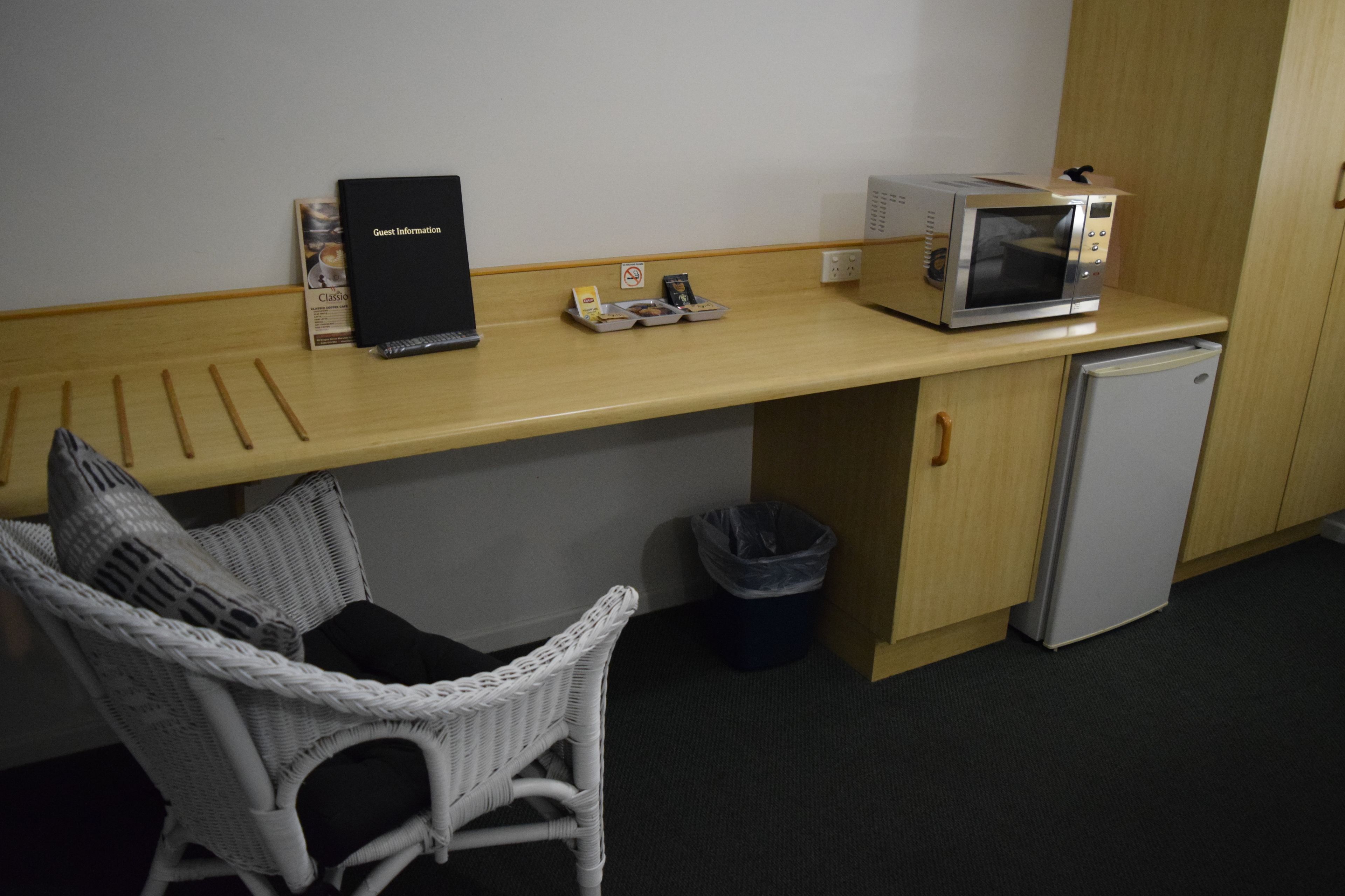 Una kitchenette con microondas, un refrigerador pequeño, una silla de mimbre, un cesto de basura y un folleto informativo para huéspedes.