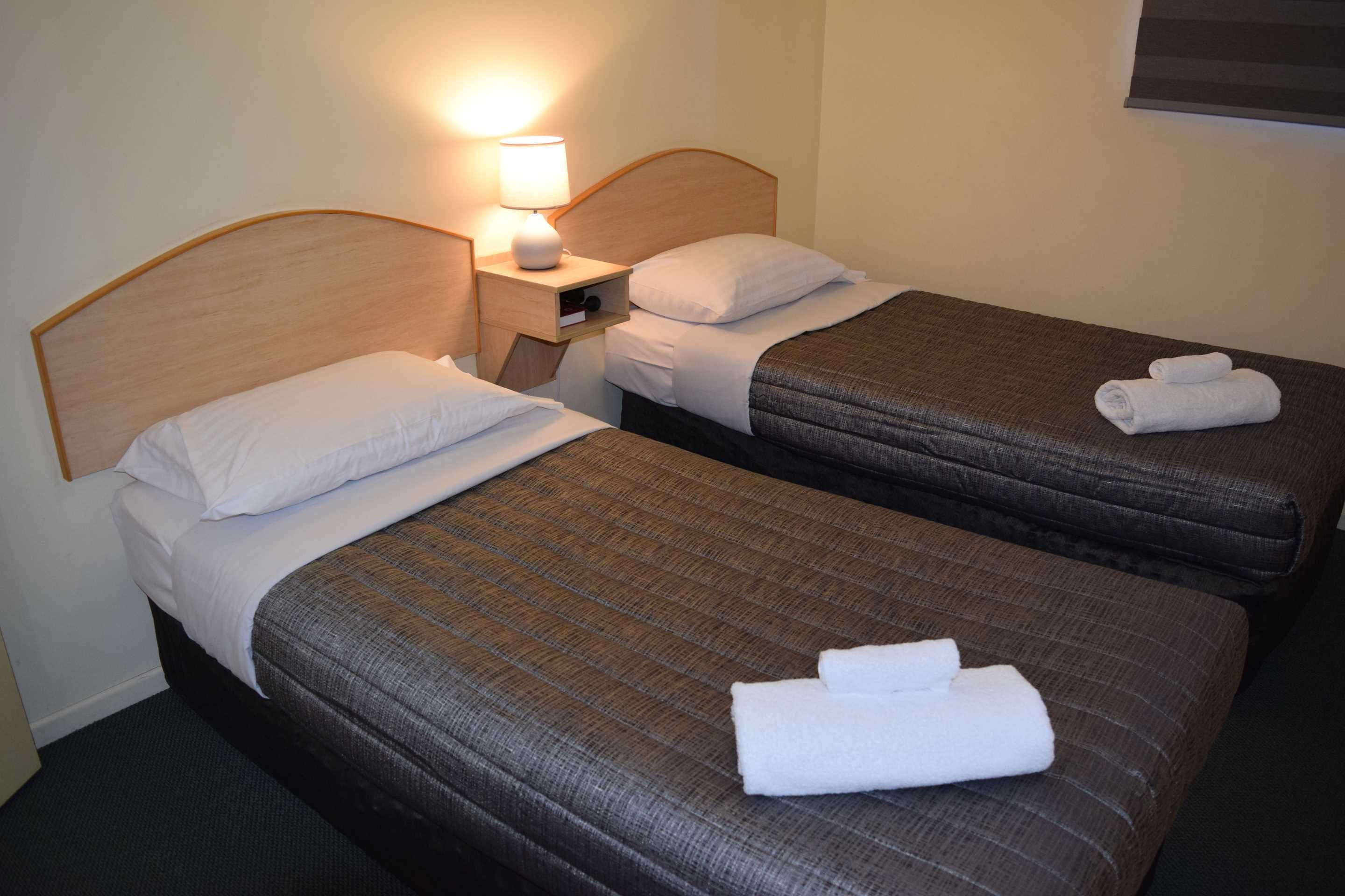 Habitación de hotel con dos camas individuales, cada una con una almohada y una toalla enrollada. Hay una mesita de noche con una lámpara entre las camas.