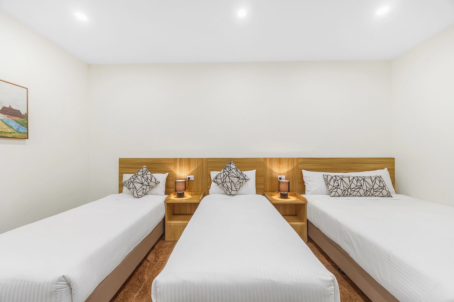 A hotel room with three single beds, each with a wooden headboard and a bedside table with a lamp.