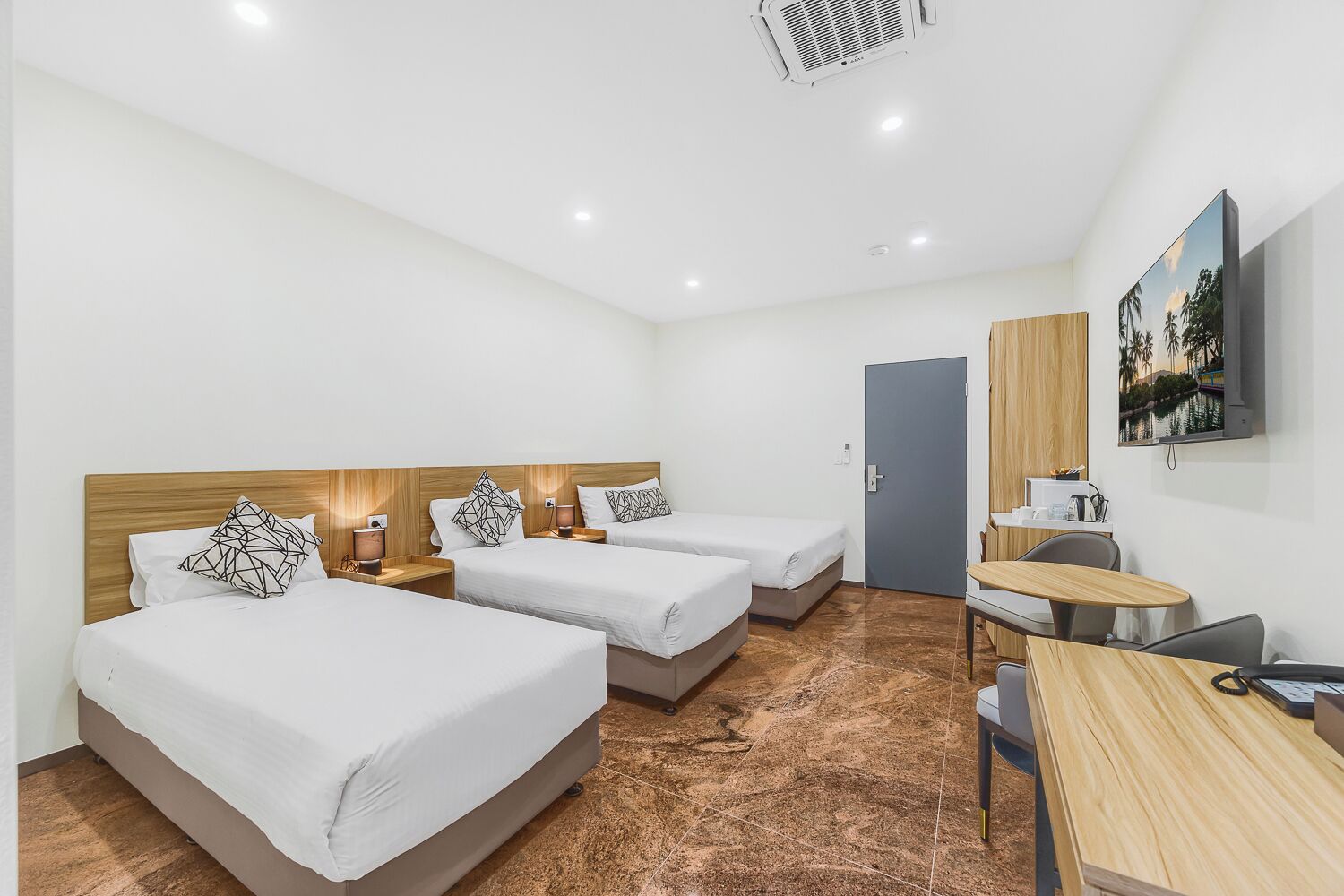 Quarto de hotel com três camas, uma escrivaninha, uma cadeira, uma televisão e uma porta.