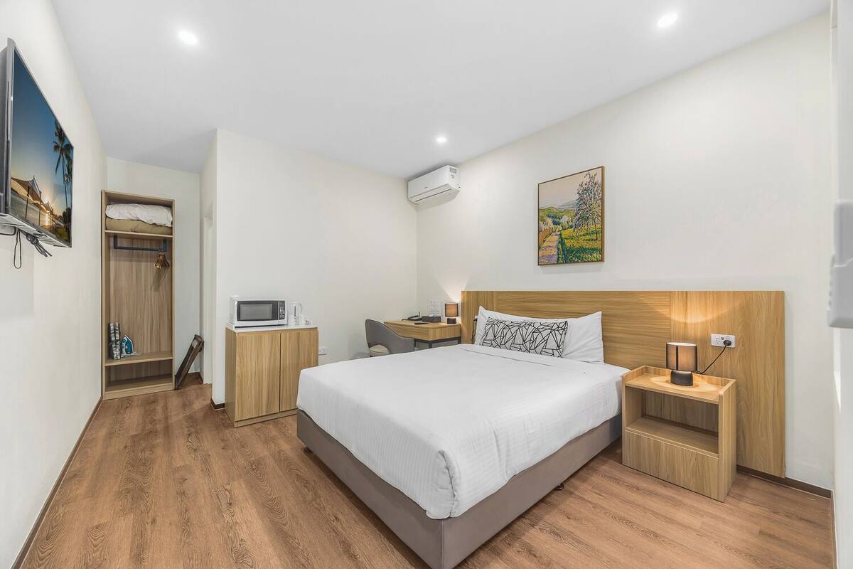 A modern hotel room with a large bed, wooden flooring, a desk with a chair, a TV, and an air conditioning unit.