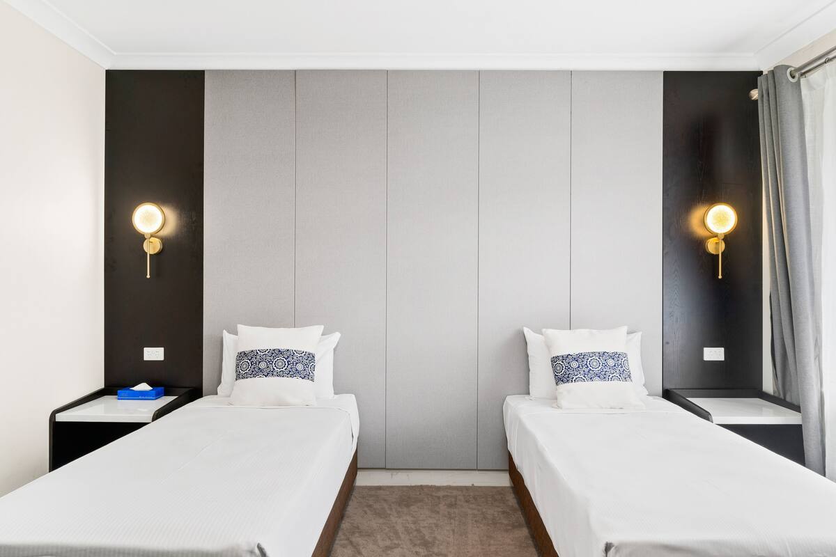 A hotel room with two single beds, a neutral color scheme, and wall-mounted light fixtures.