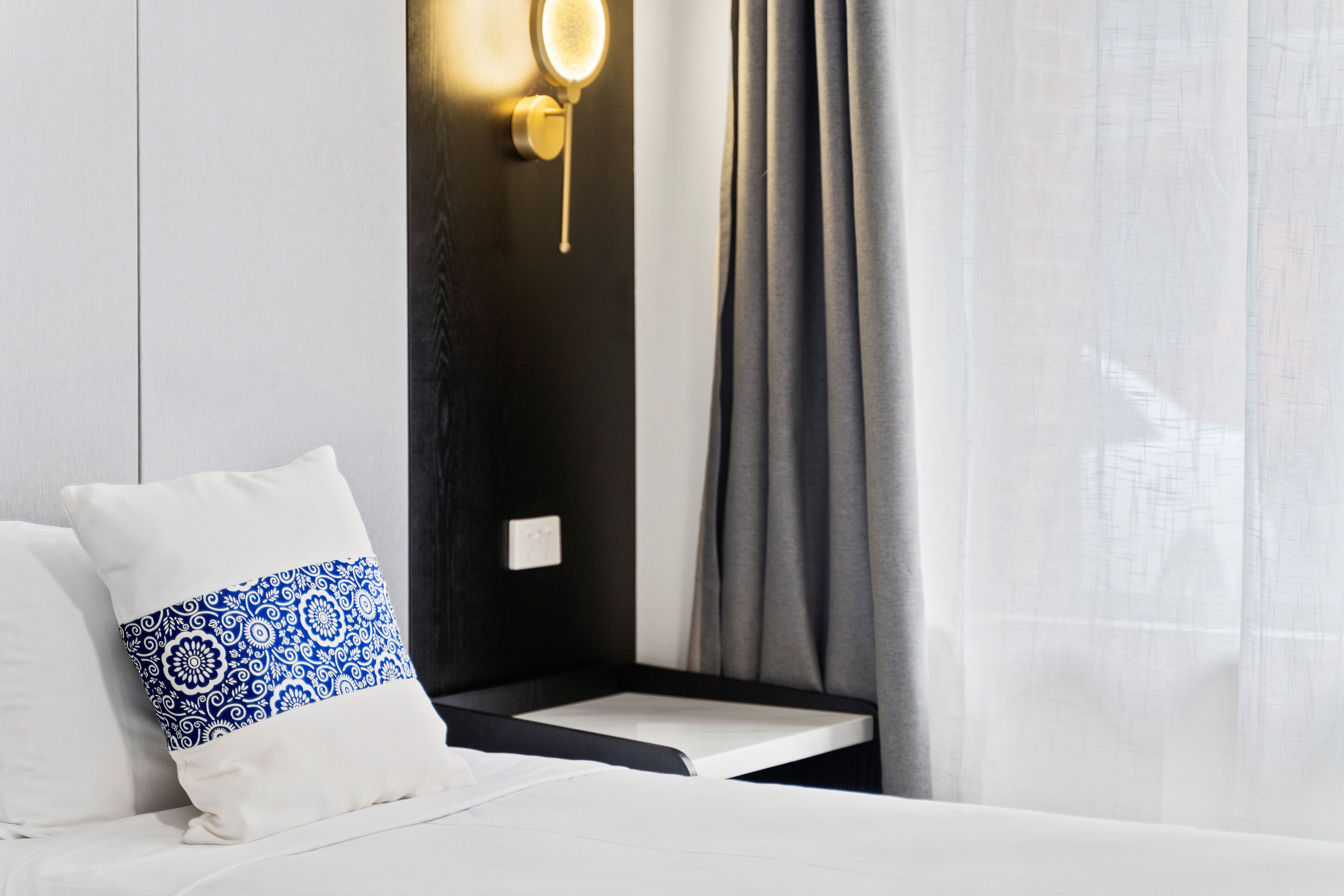 A modern hotel room with a bed, a bedside table, and a wall-mounted light fixture.