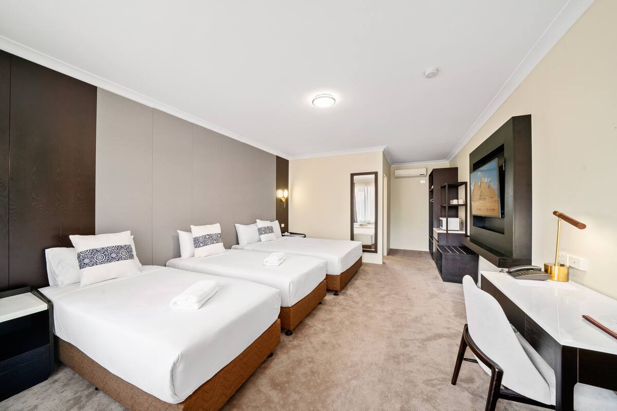 A hotel room with three beds, a desk, a TV, and a bathroom visible through an open door.