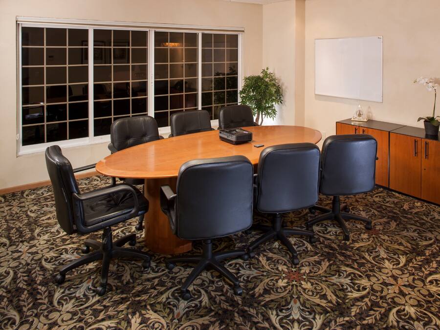 Un salón de conferencias con una gran mesa ovalada, seis sillas, un proyector, una pizarra, una planta en maceta y un teléfono sobre un mueble.