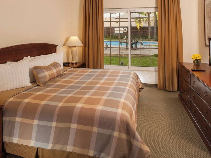 Habitación de hotel con cama, televisor, cómoda, lámpara y vistas a una piscina y zona exterior.