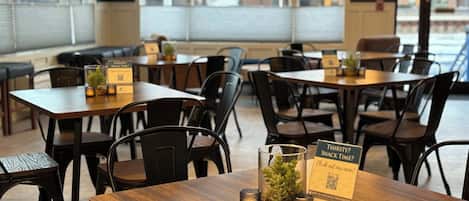 L’intérieur d’un restaurant, avec des tables en bois et des chaises noires, où l’on trouve des codes QR pour accéder aux menus numériques et un panneau indiquant la possibilité de commander des boissons non alcoolisées.
