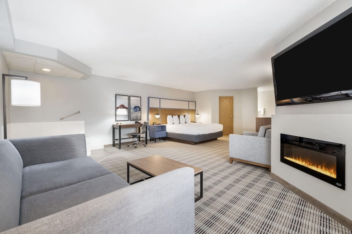 A modern hotel room with a large bed, a desk, a sofa, and a fireplace.