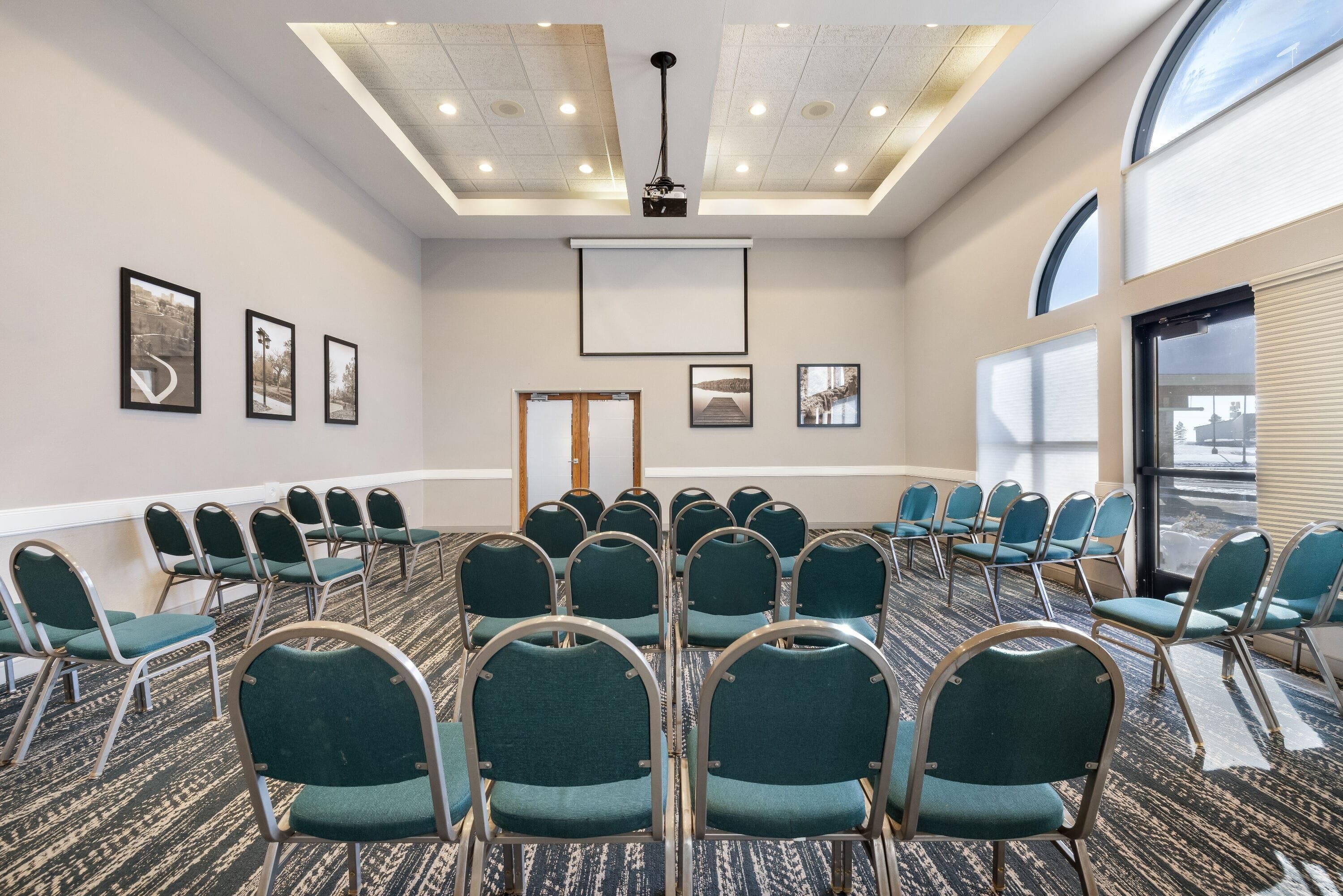 A conference room with rows of teal chairs, a projector screen, and framed pictures on the walls.