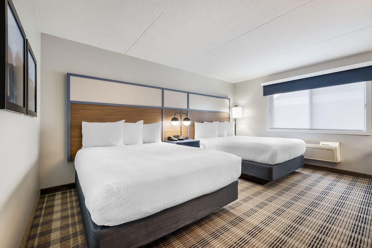 A hotel room with two beds, a headboard with built-in lighting, a bedside table with a lamp, and a large window providing natural light.