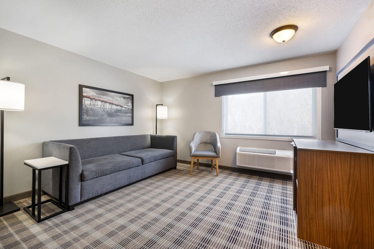 A modern hotel room with a grey sofa, a small side table, a chair, a large window with a view, and a flat-screen TV.