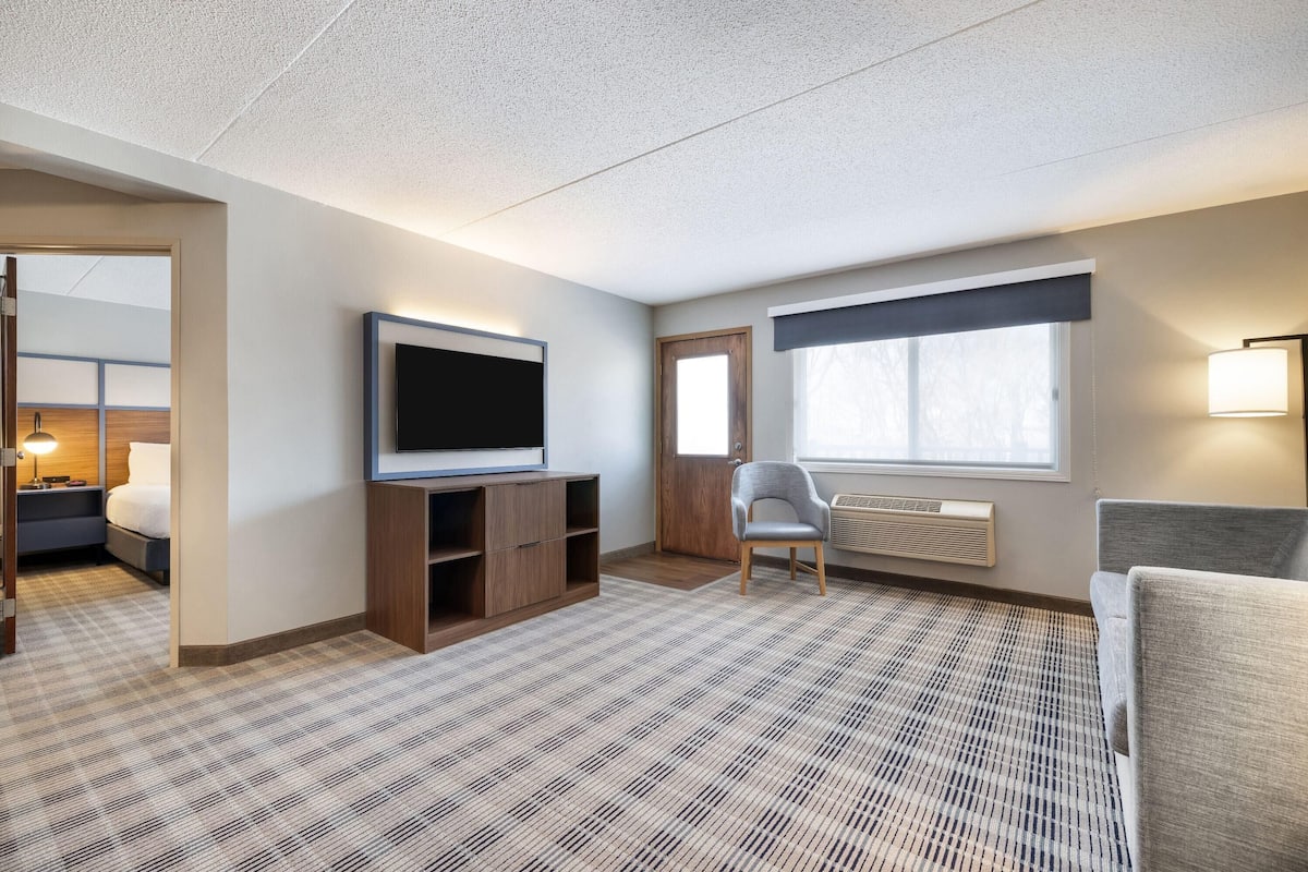 A hotel room with a large flat-screen TV, a seating area with a chair, a sofa, and a window with a view.