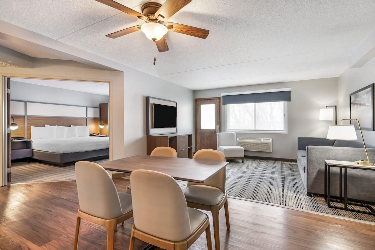 A hotel room with a bed, a TV, a desk with chairs, a sofa, and a ceiling fan.
