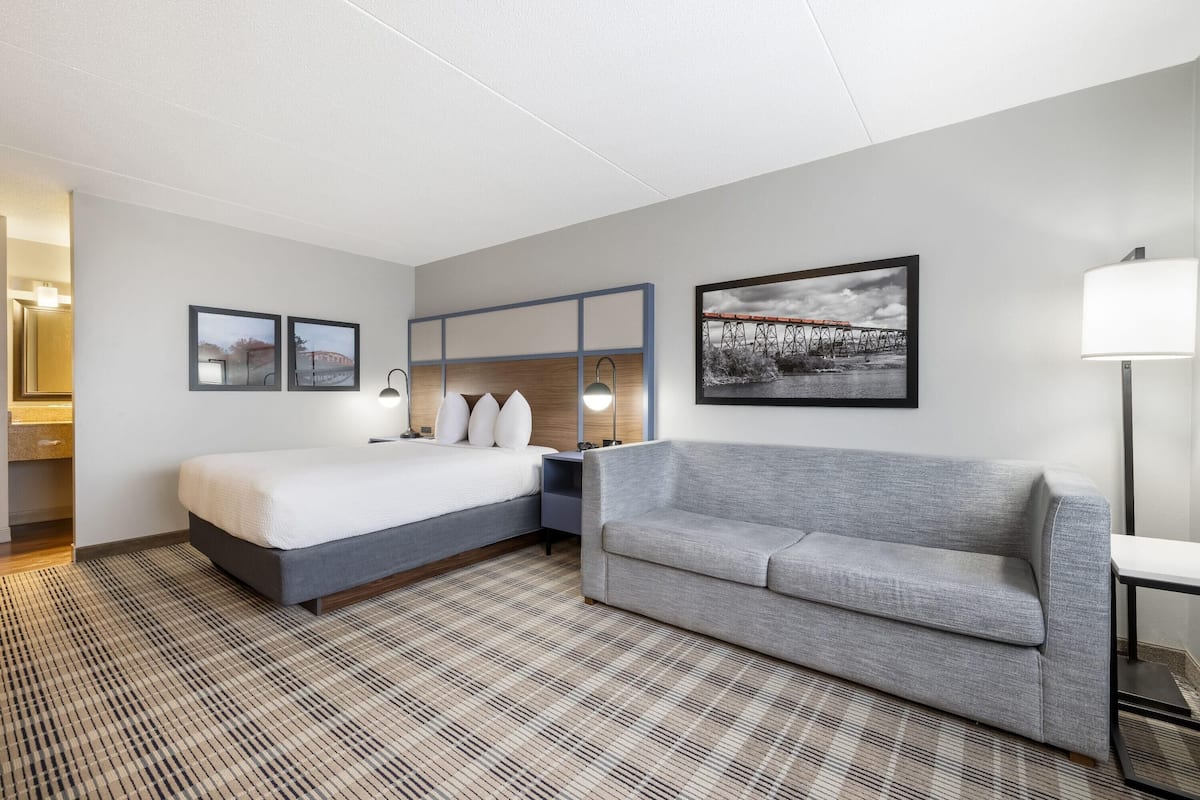 A hotel room with a bed, a gray sofa, a wall-mounted TV, and a framed picture of a bridge.