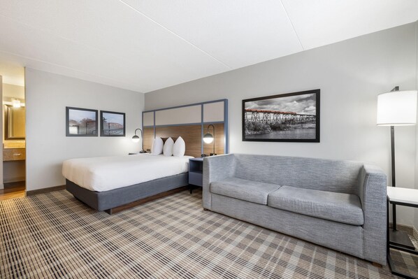 Una habitación de hotel con una cama, un sofá gris, un televisor montado en la pared y un cuadro con una imagen de un puente.