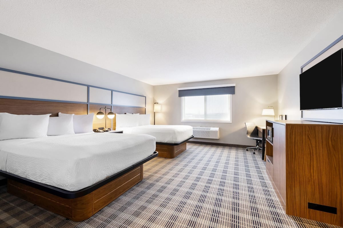 A hotel room with two beds, a desk, a flat-screen TV, and a window with blinds.