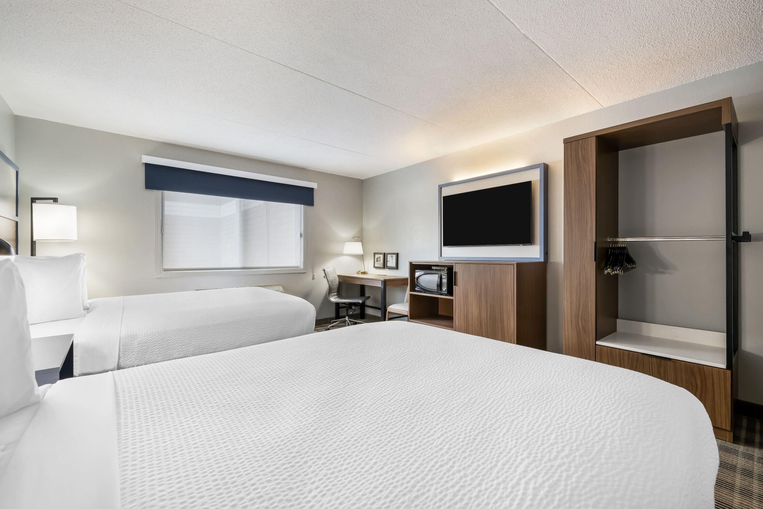 Ein Hotelzimmer mit zwei Betten, einem Schreibtisch mit Stuhl, einem Fernseher, einem Kleiderschrank und einem kleinen Tisch mit Lampe.