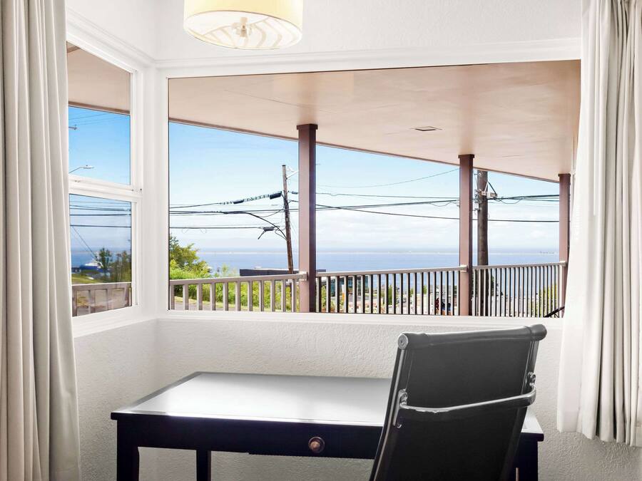 A room with a desk and chair, large windows with curtains, a view of the sea, and a balcony with a railing.