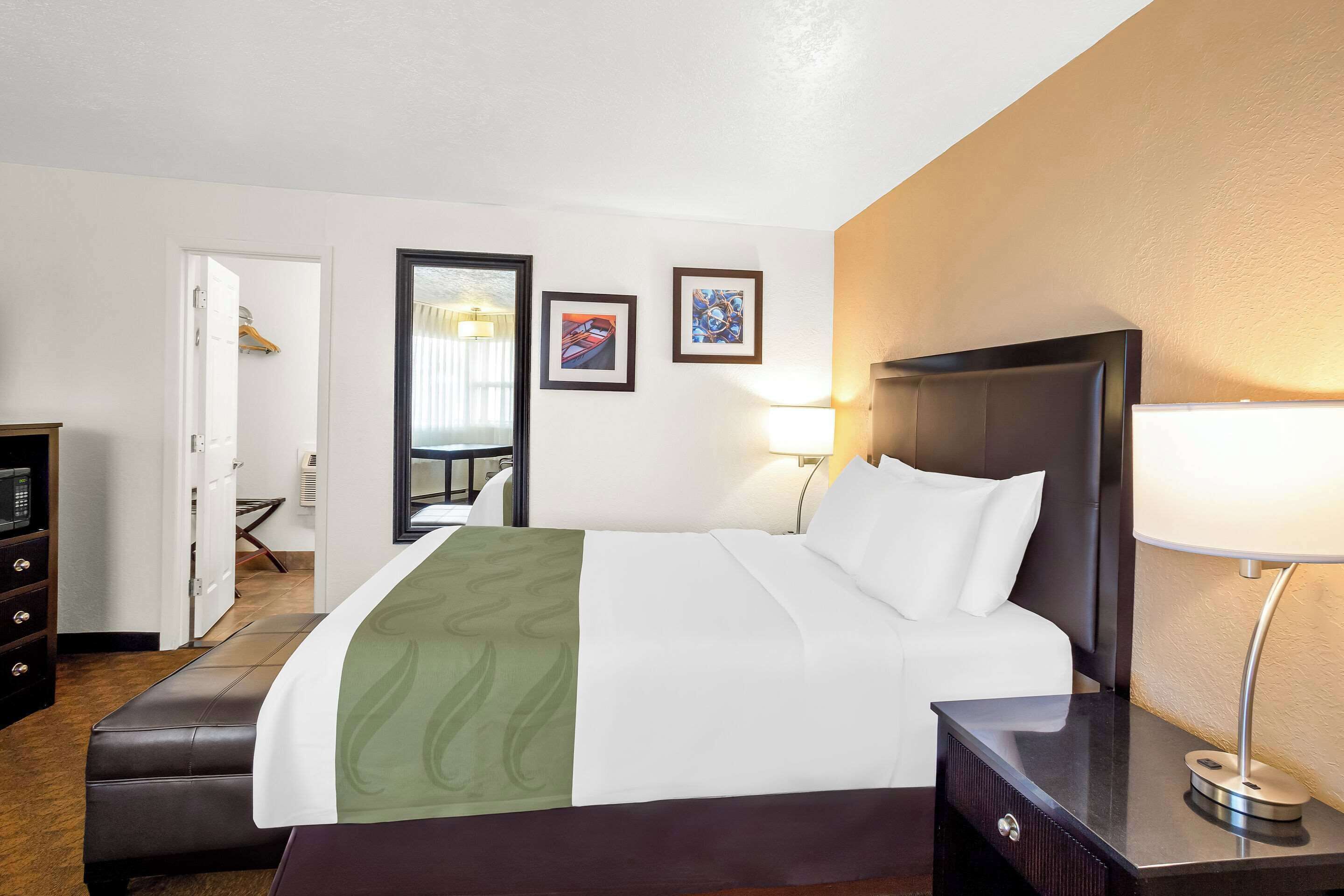 Habitación de hotel con cama, mesita de noche, lámpara y vista al pasillo.