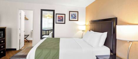 Habitación de hotel con cama, mesita de noche, lámpara y vista al pasillo.