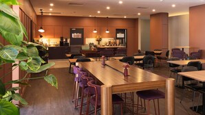 A modern dining area with wooden tables and purple bar stools, a kitchen in the background, and ceiling lights. - Mercure Newcastle (Newcastle West)