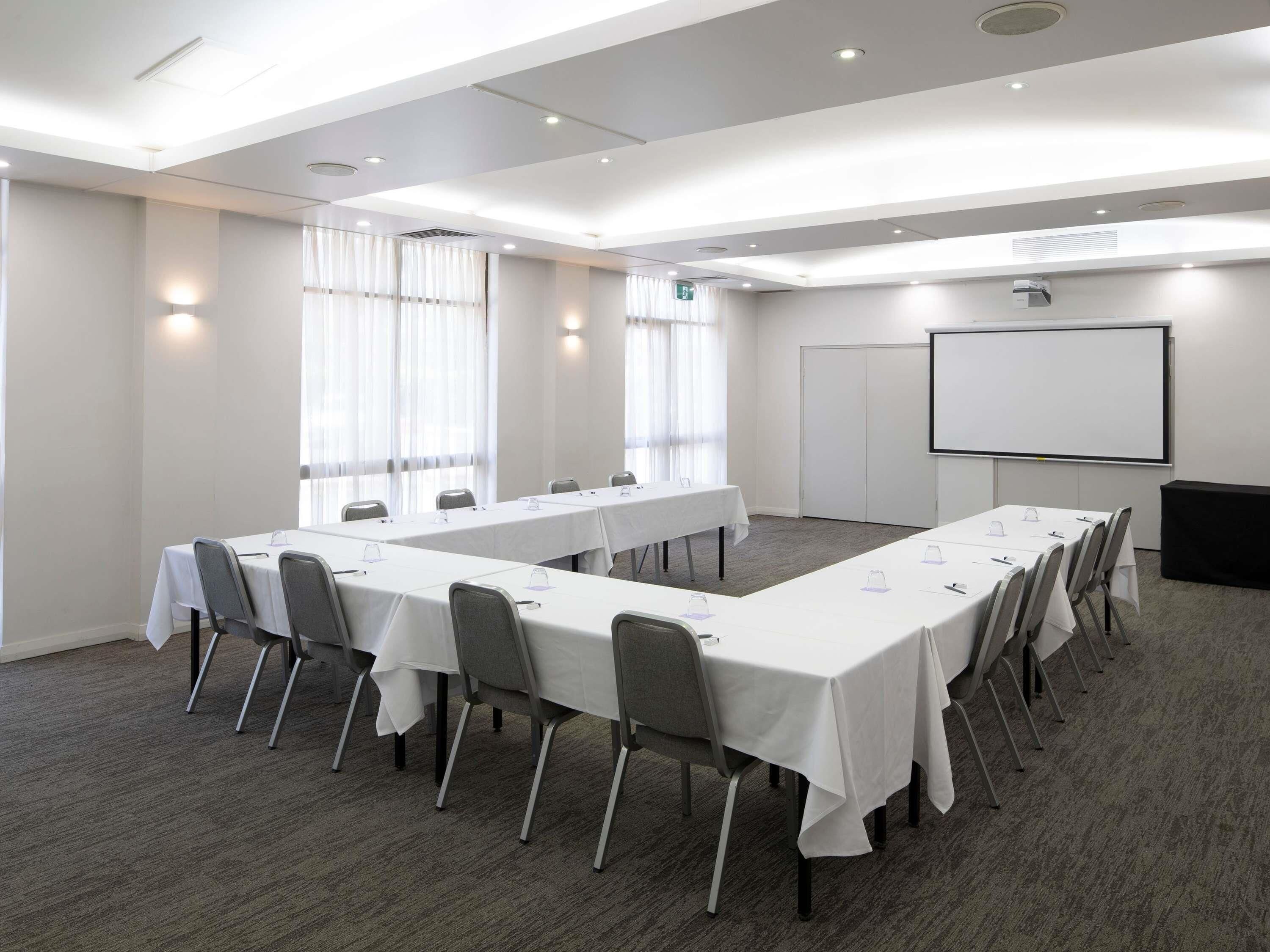 Ein Konferenzraum mit langen Tischen, Stühlen, einer Projektionsleinwand und einer Whiteboard-Wand.