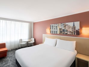 A hotel room with a bed, a chair, a small table, and a wall-mounted lamp. - Mercure Newcastle (Newcastle West)