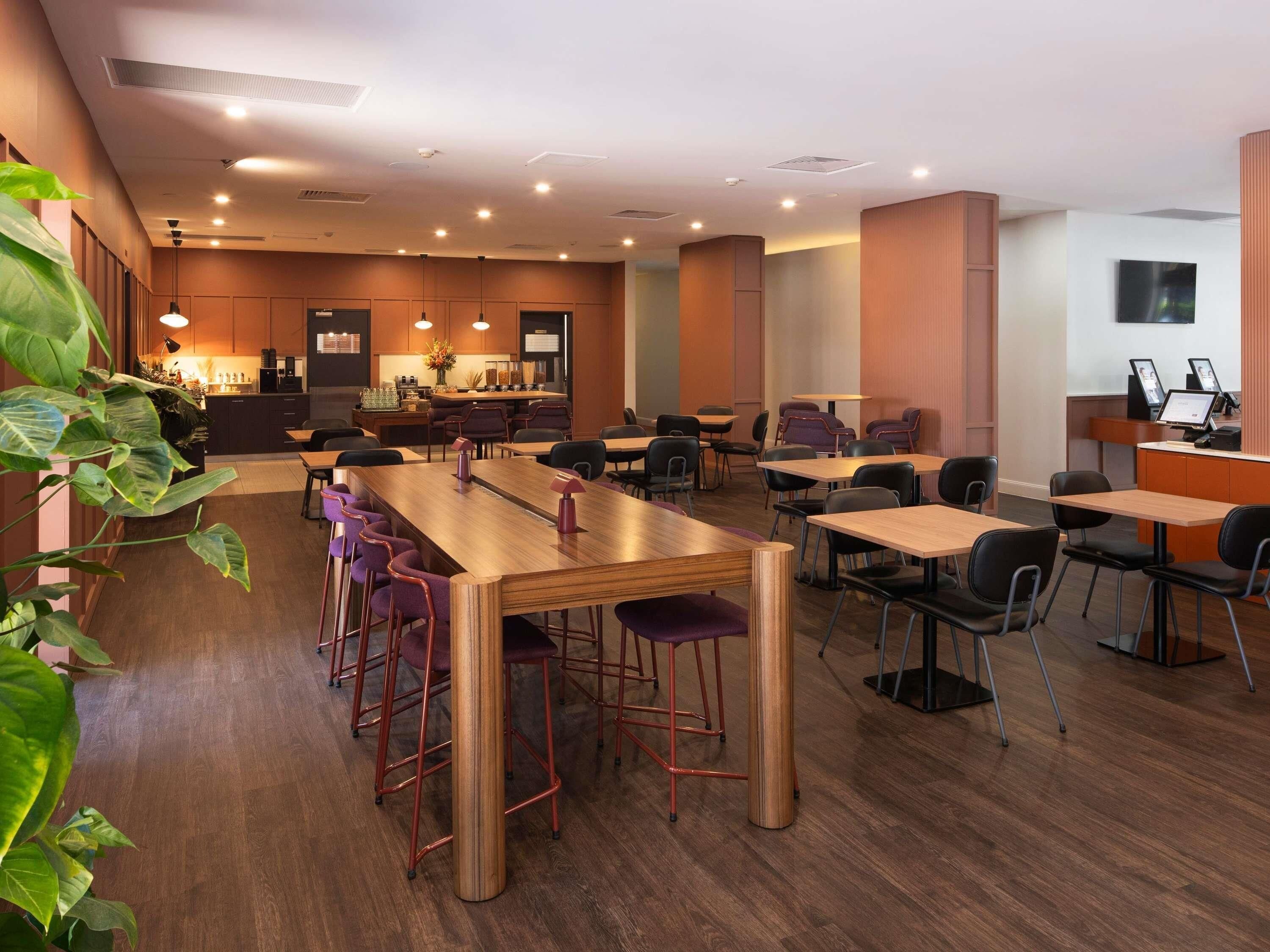 A modern restaurant with a long wooden table, purple bar stools, and a bar area in the background.