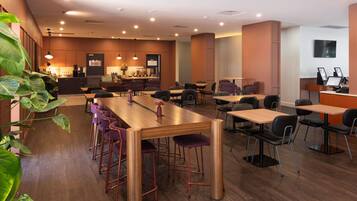 A modern restaurant with a long wooden table, purple bar stools, and a bar area in the background.