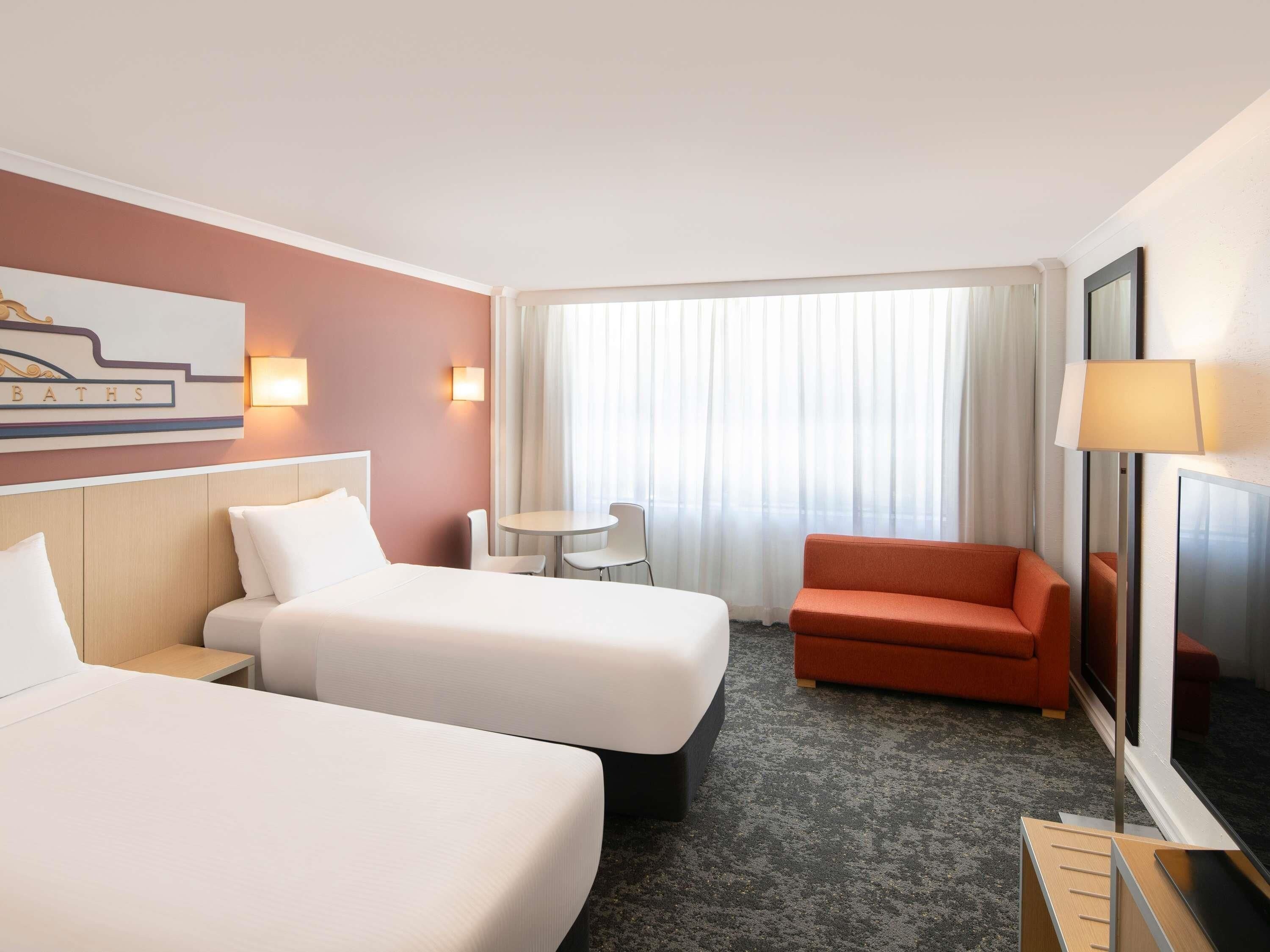 A hotel room with two beds, a small table, a sofa, a TV, and a window with curtains.